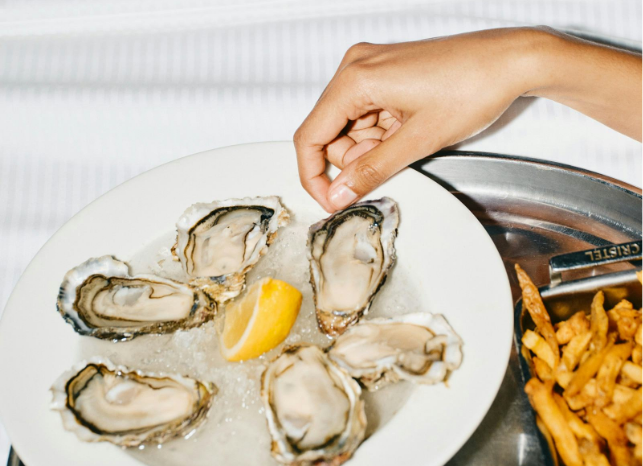 Woman reaches for an oyster. Oysters lie on a bed of ice and next to them is a portion of French fries.