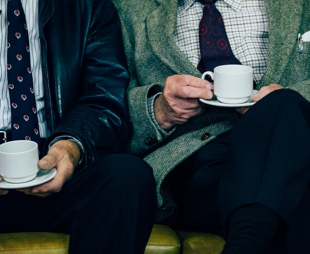 Two older men sit next to each other. One of them is drinking a coffee.